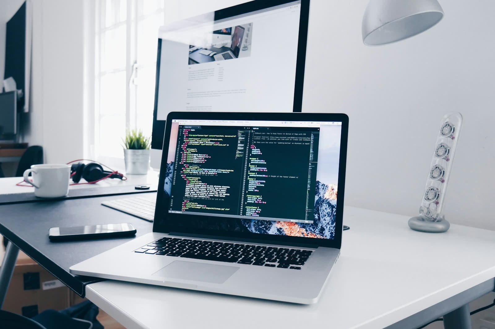 A laptop on a desk with a code editor open, a second monitor showing the page being worked on