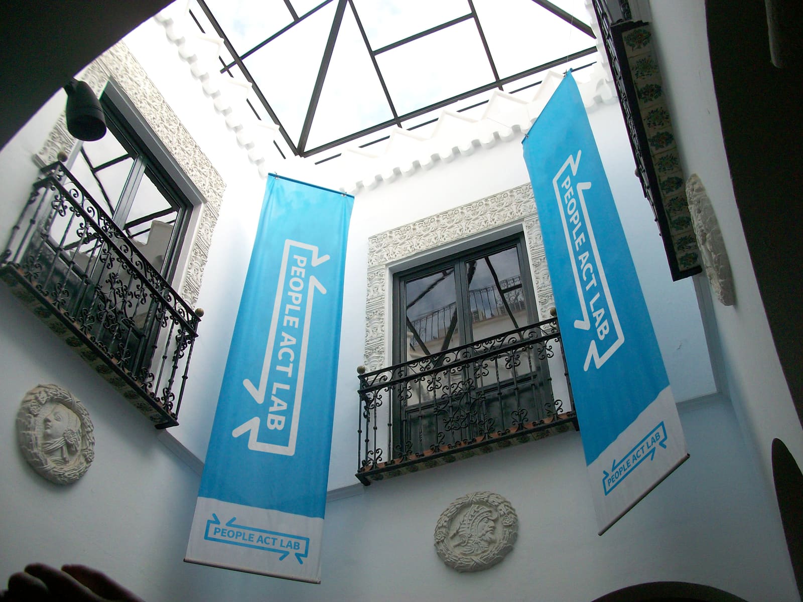 Two People Act Lab banners hanging in a white skylit courtyard, the wordmark bracketed by inward arrows on cyan.
